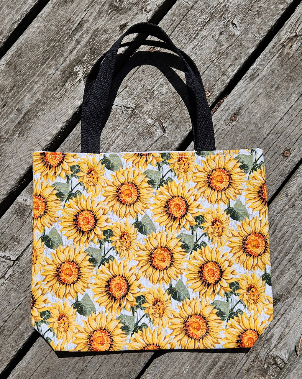 A tote bag with a vibrant sunflower print and black handles, resting on a weathered gray wooden plank surface.