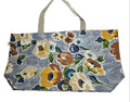 A fabric tote bag with a floral print on a dusty blue-gray background, featuring cream, yellow, brown, and blue flowers with green leaves. The bag has a light beige canvas handle and is laid flat on a white fabric surface.