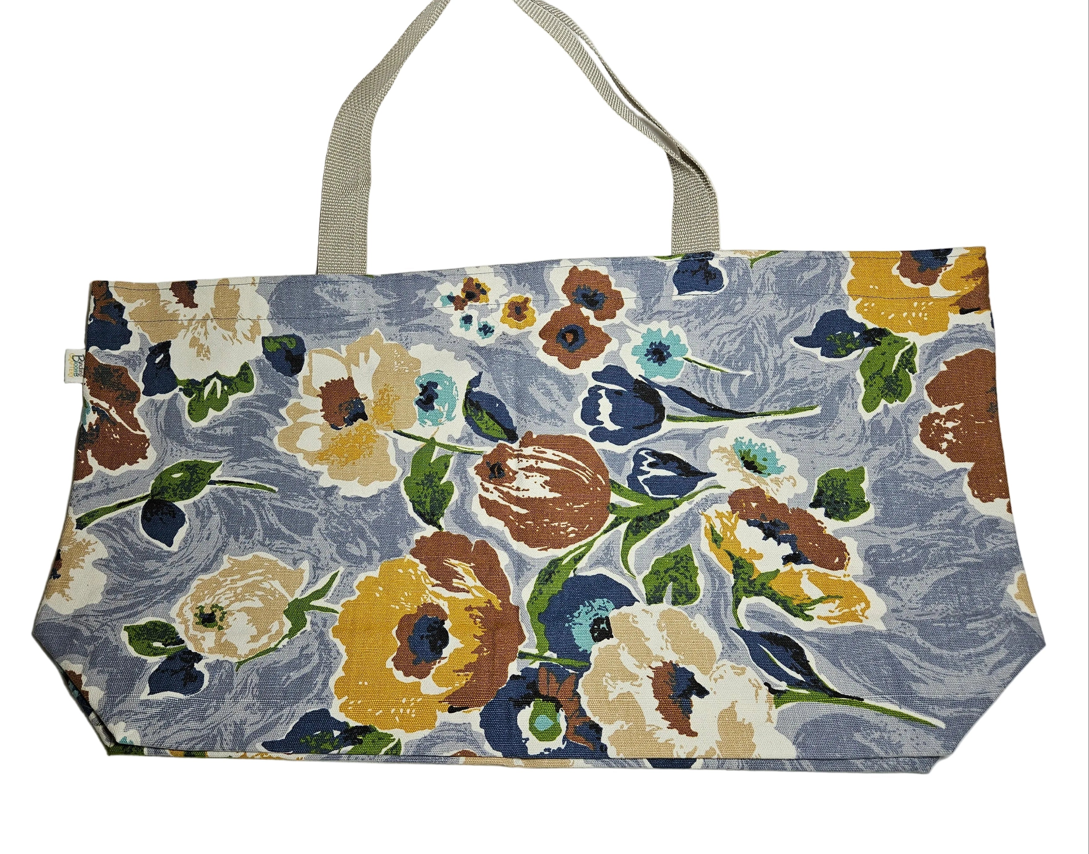 A fabric tote bag with a floral print on a dusty blue-gray background, featuring cream, yellow, brown, and blue flowers with green leaves. The bag has a light beige canvas handle and is laid flat on a white fabric surface.